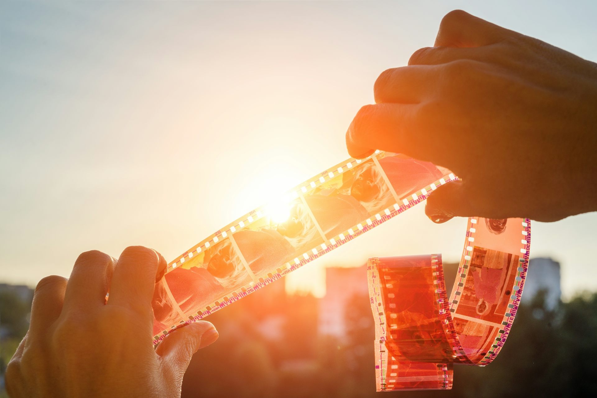 Close-up of two hands holding a strip of photographic negatives up against a sunset sky, warm orange and gold tones